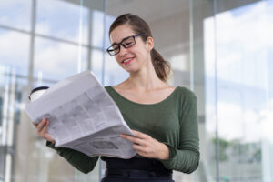 Positive business expert checking out financial news while drinking coffee outside office. Smiling young woman in eyeglasses reading newspaper. Business information concept